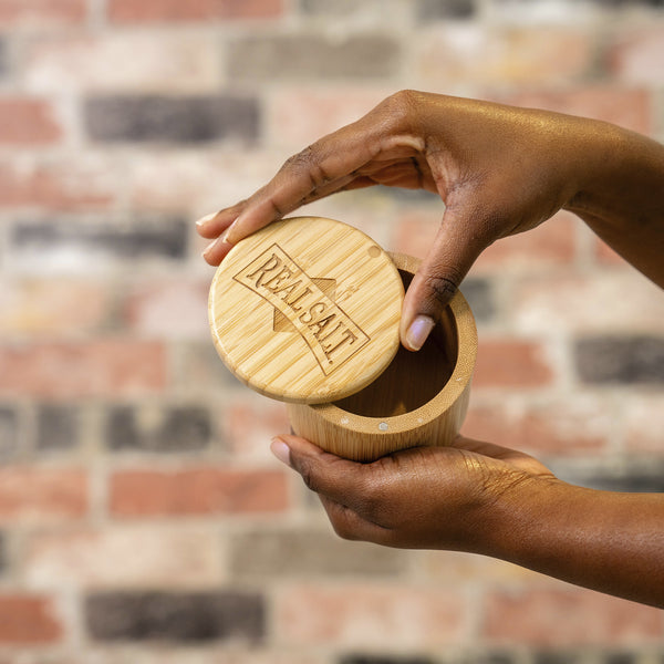 Hands lift the lid of a bamboo salt cellar revealing the "Real Salt" logo. 
