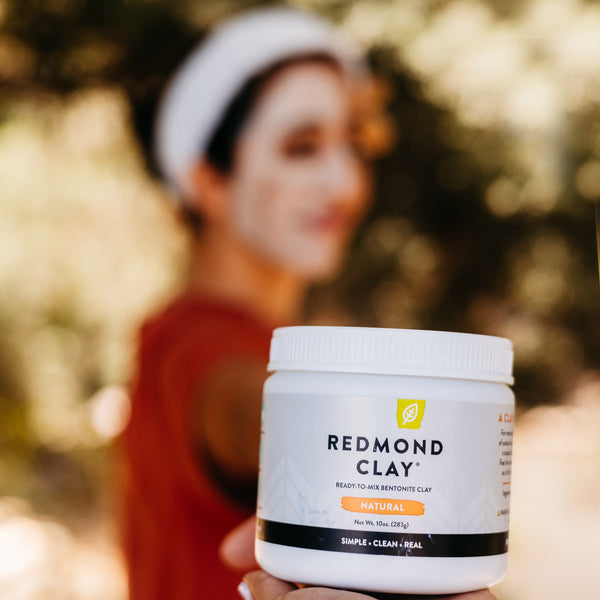 Hand holds out a container of Redmond Clay against a blurred background of a woman wearing a face mask. 
