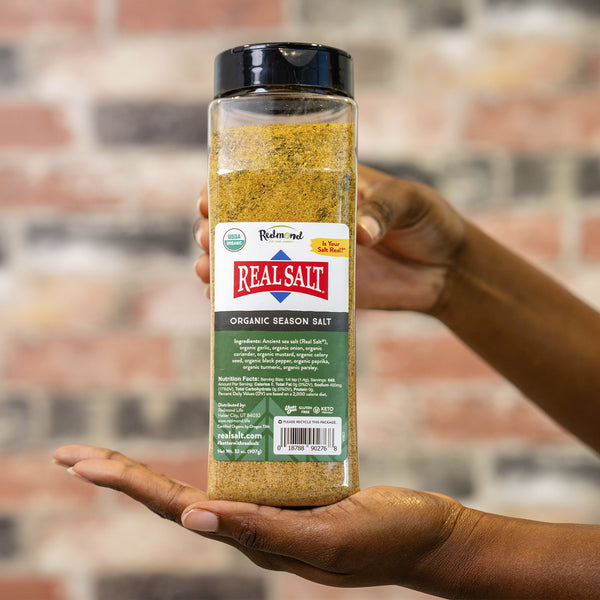 Hand holds a jar of Redmond Real Salt Organic Season Salt against a brick background. 
