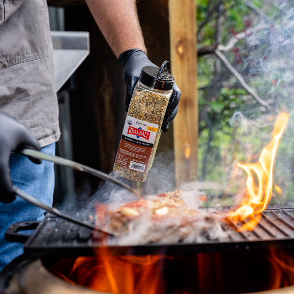 Chef sprinkles Redmond Real Salt Organic Garlic Pepper on a sizzling steak over a fiery grill. 
