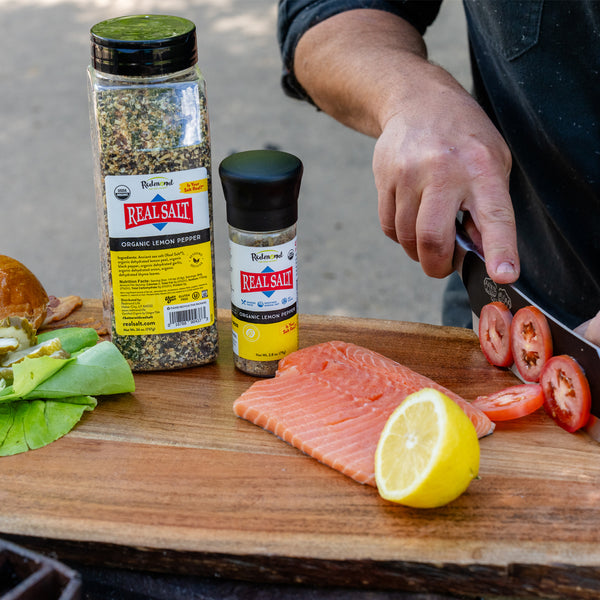 Chef slices tomatoes beside fresh salmon and a lemon wedge seasoned with Redmond Real Salt Organic Lemon Pepper. 
