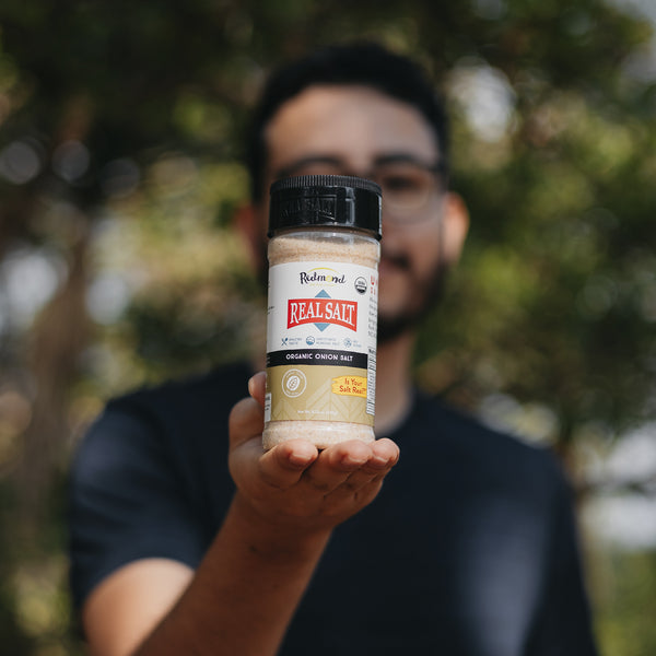 Man holds out a shaker of Redmond Real Salt Organic Onion Salt.