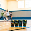 Four Re-Lyte Hydration 30 count stick packs electrolyte powder on a basketball court with young men playing basketball in the background. Flavors from left to right: Strawberry Lemonade, Mango, Unflavored and Piña Colada.