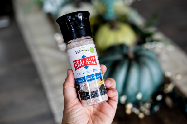 Hand holds a grinder of coarse Redmond Real Salt against a blurred background.