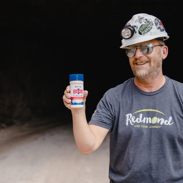 Miner wearing headlamp and sunglasses holds a container of Redmond Real Salt. 

