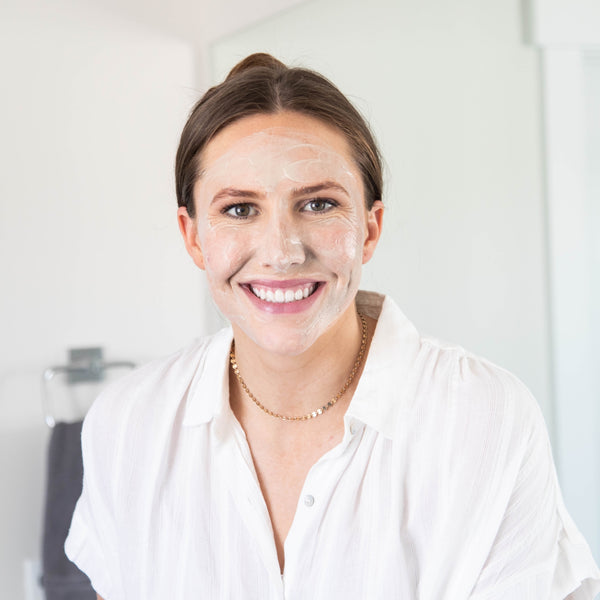 Woman smiles at the camera wearing a white skincare face mask. 
